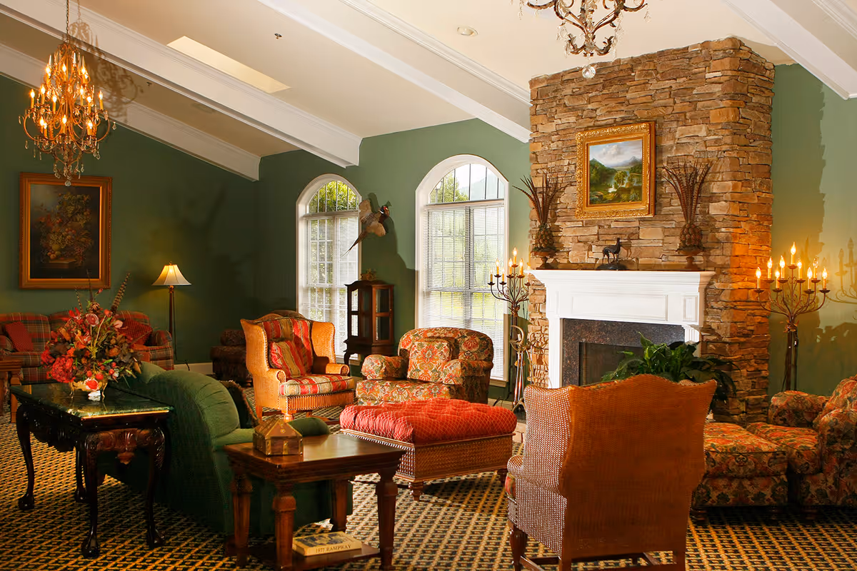 A cozy senior living facility lounge with green walls, a stone fireplace, and multiple upholstered chairs and sofas arranged around a coffee table. The room features large arched windows, chandeliers, decorative lamps, floral arrangements, and framed artwork on the walls.