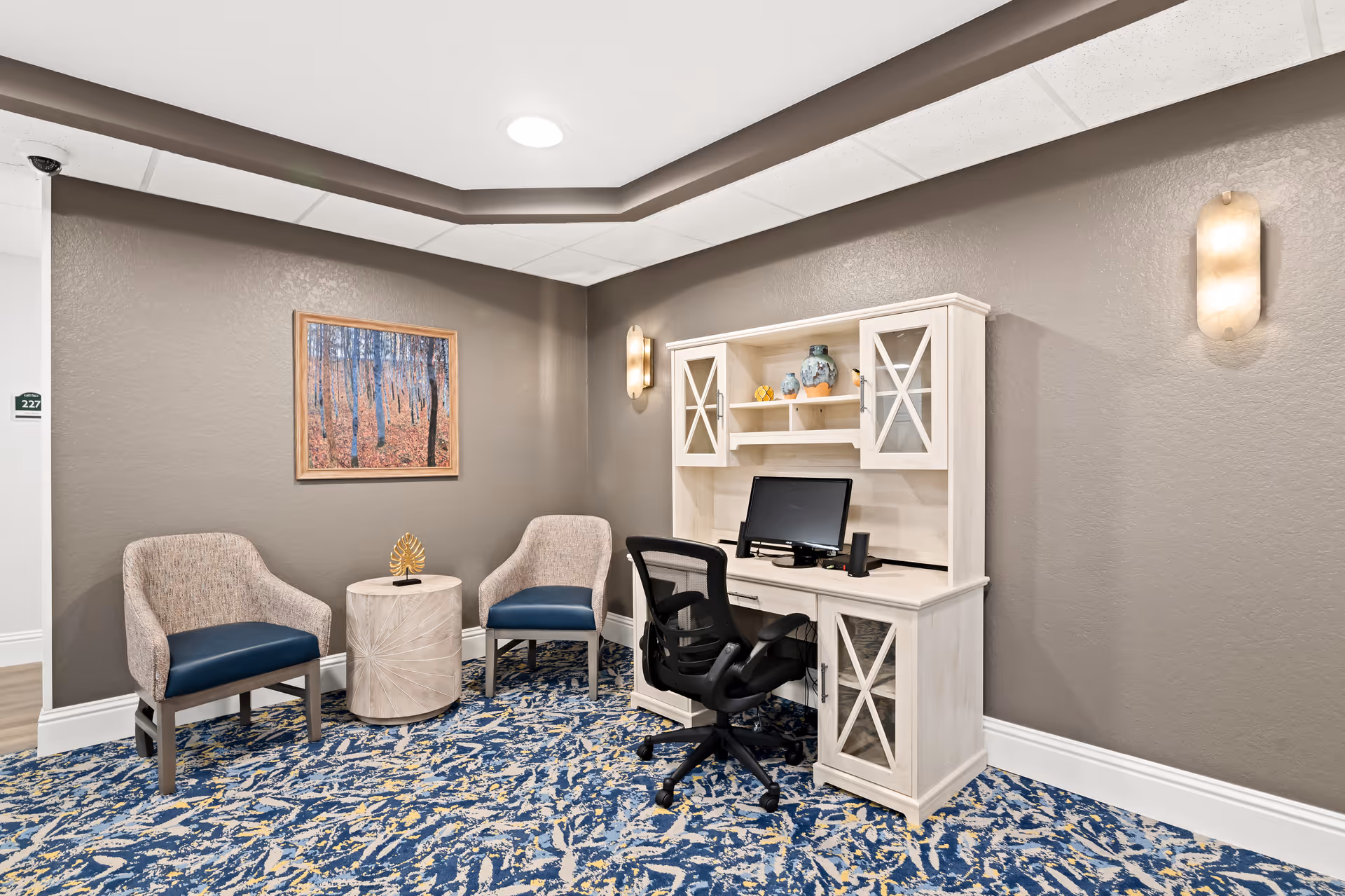 A small office area in a senior living facility with a white wooden desk and hutch holding a computer monitor and decorative items. There is a black office chair in front of the desk, and two upholstered chairs with blue cushions flank a small round table. The walls are painted taupe, with a framed picture of a forest hanging on one wall and two wall sconces providing light. The floor is covered with a blue and beige patterned carpet.