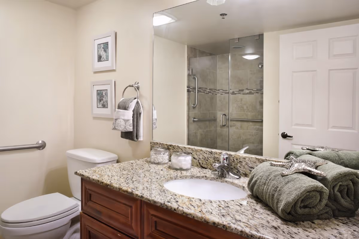 A clean bathroom with a granite vanity and sink, rolled green towels, toilet, and a glass-enclosed shower reflected in the mirror.