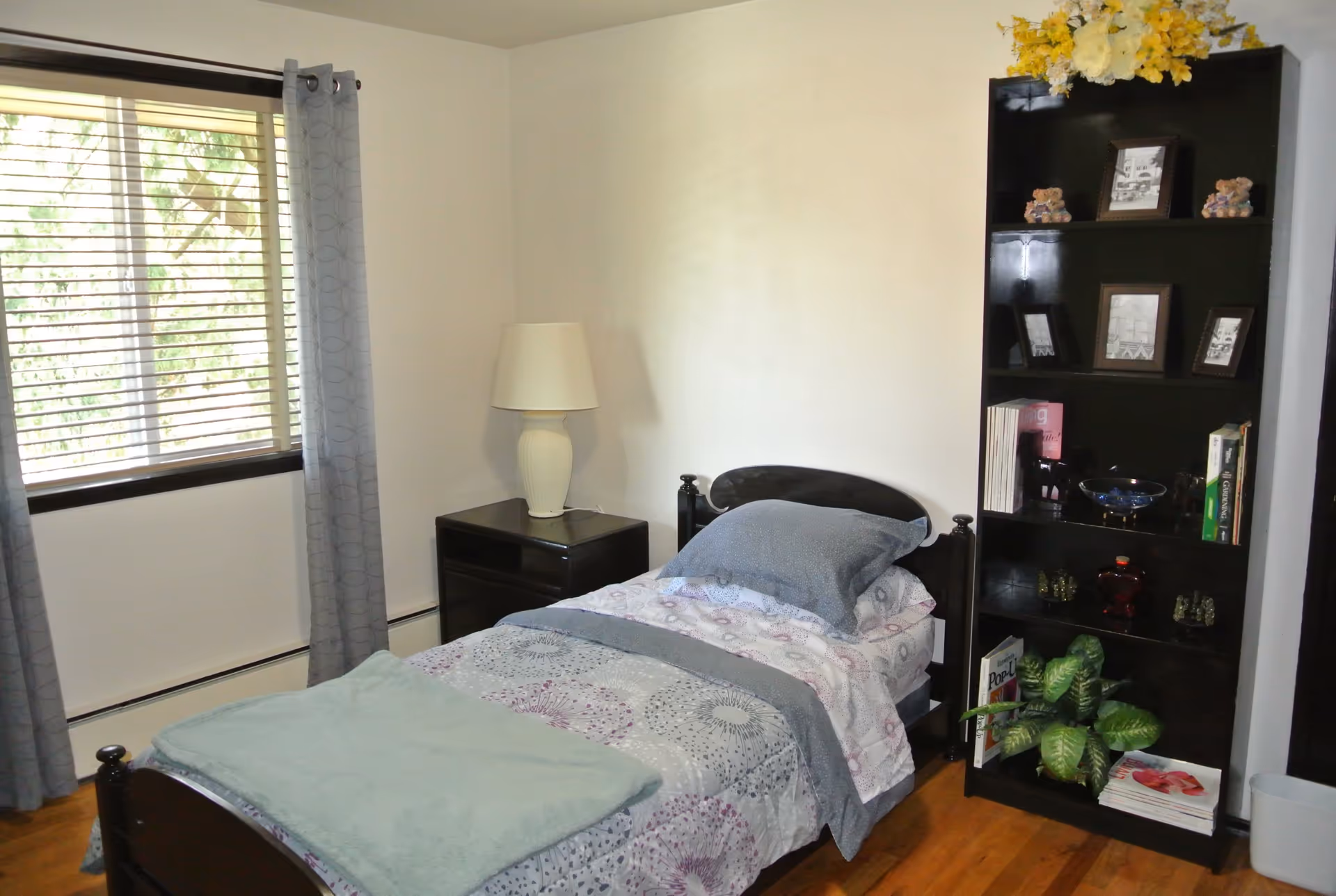 A small bedroom with a single bed made up with patterned bedding and a folded blanket at the foot. Next to the bed is a dark wooden nightstand with a white lamp. A tall dark wooden bookshelf stands against the wall, decorated with framed photos, small teddy bears, books, a plant, and other decorative items. A window with blinds and gray curtains is on the left side, letting in natural light.