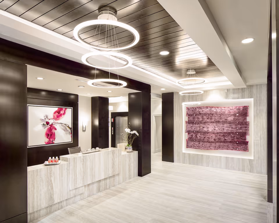 Modern reception area with a light-colored marble desk, black pillars, and contemporary circular ceiling lights. The walls feature abstract artwork in shades of pink and purple. The floor is tiled in light gray, and there is a small vase with white flowers on the desk.