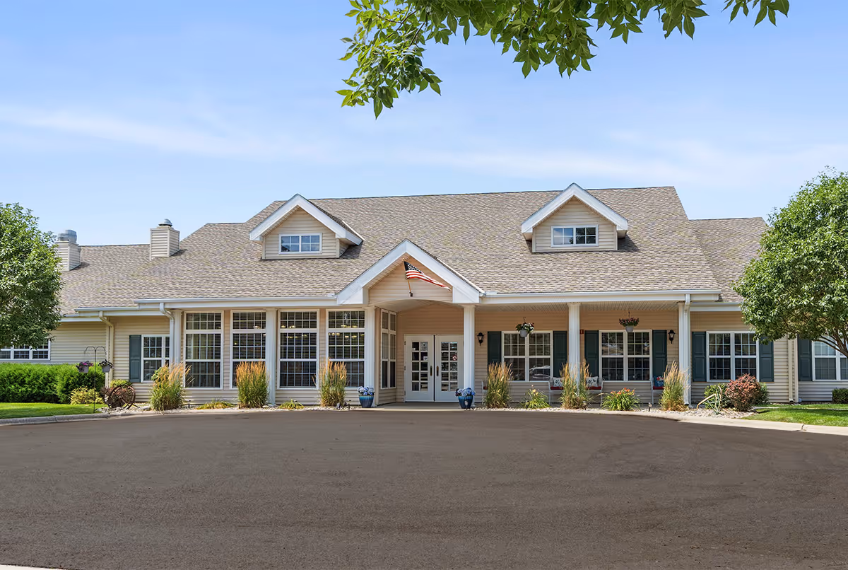 Front exterior view of a single-story senior living facility building with a pitched roof, multiple windows, an American flag above the entrance, and landscaping with trees and shrubs under a clear blue sky.