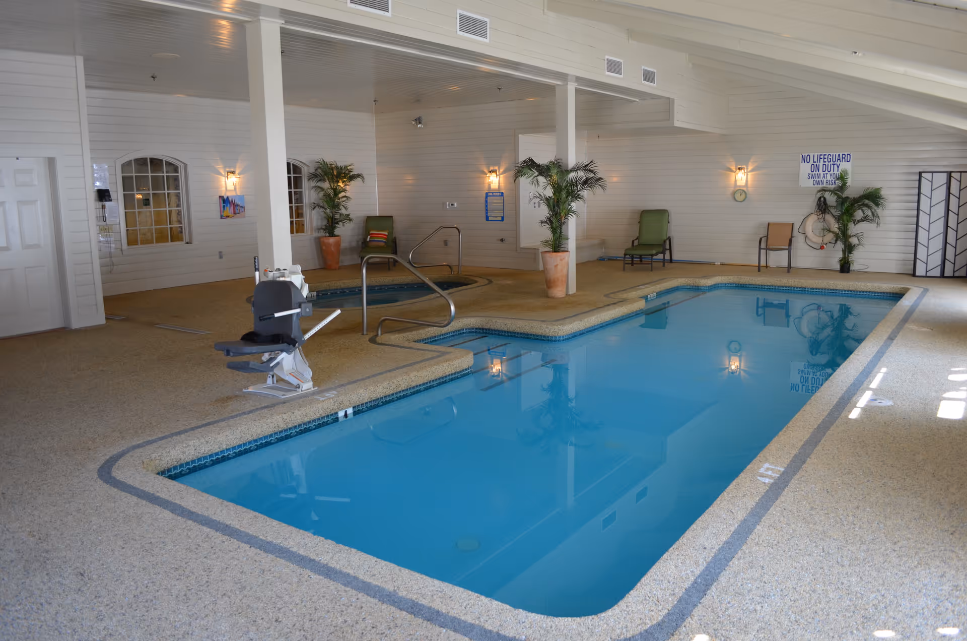 Indoor swimming pool and hot tub area with a pool lift, seating, and potted plants in a senior living facility.