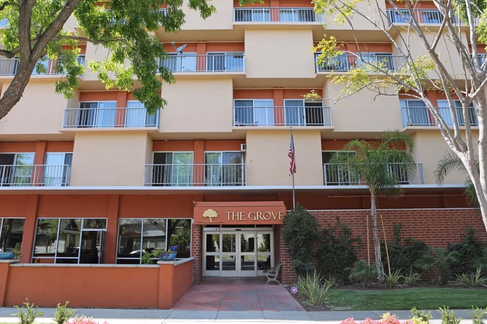 Front exterior view of The Grove Care and Wellness building with multiple balconies, an entrance with glass doors, an American flag, and surrounding trees and plants.
