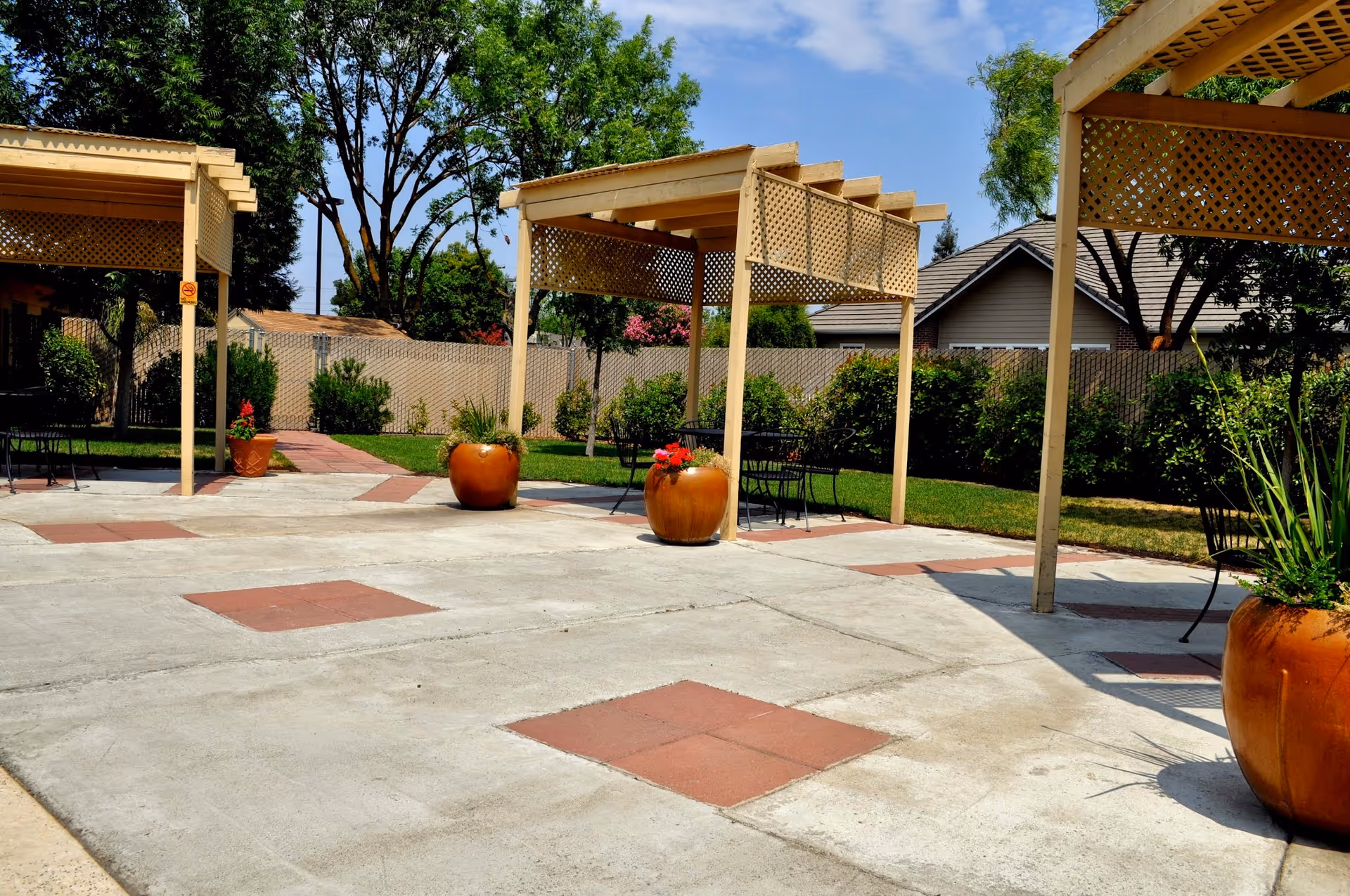 Outdoor patio area with concrete flooring featuring red square tiles, three beige pergolas with lattice sides, large orange planters with green plants and flowers, metal chairs and tables, surrounded by green grass, bushes, trees, and a fence with houses visible in the background under a blue sky with some clouds.