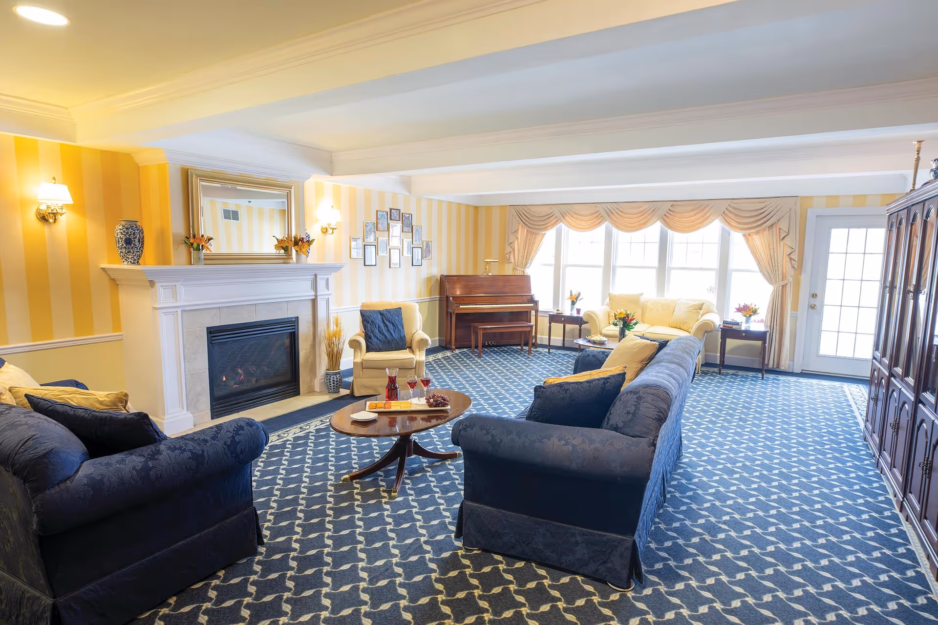 A bright and spacious living room with blue patterned carpet, yellow striped walls, and white ceiling beams. The room features a fireplace with a mirror above it, two blue sofas, two beige armchairs, a wooden coffee table with drinks and snacks, a piano against the far wall, and large windows with beige curtains letting in natural light.