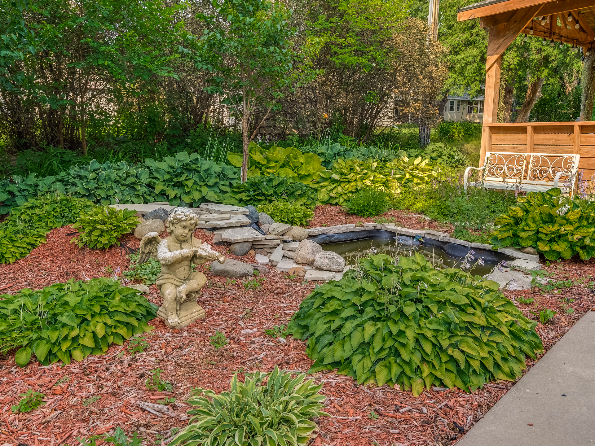 A garden area with lush green plants and shrubs surrounding a small pond with rocks. There is a stone statue of a cherub playing a violin in the garden. To the right, there is a wooden gazebo with a white ornate metal bench underneath. Trees and additional greenery are visible in the background.