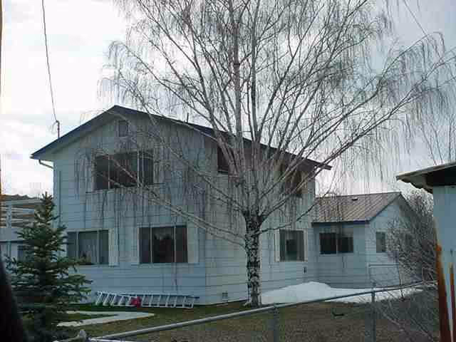 Exterior view of a two-story white building with multiple windows, a leafless tree in front, and a small patch of snow on the ground. A ladder is lying on the grass near the building, and a chain-link fence is visible in the foreground.
