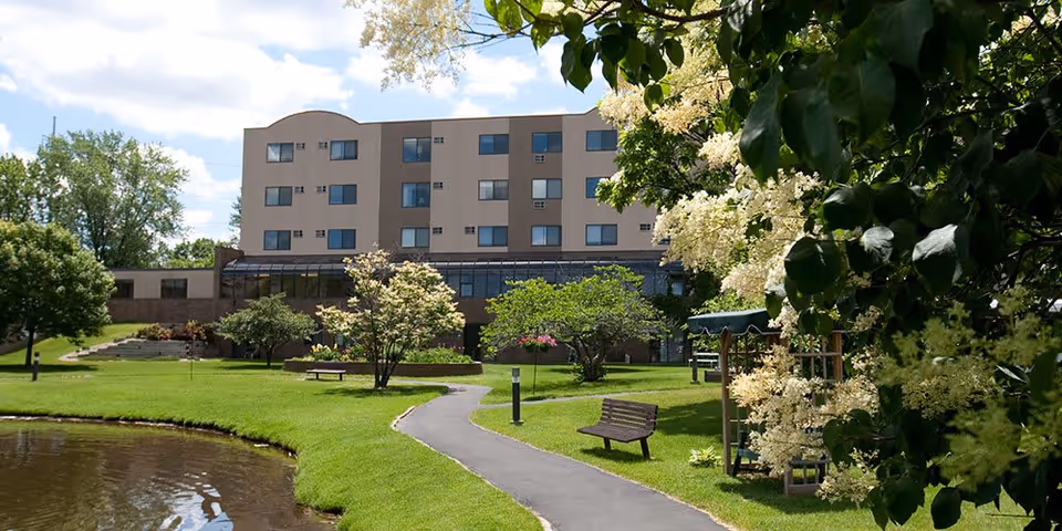 A senior living facility named Chandler Place with a multi-story building in the background, surrounded by a well-maintained garden featuring a walking path, benches, trees, flowering bushes, and a small pond under a partly cloudy sky.