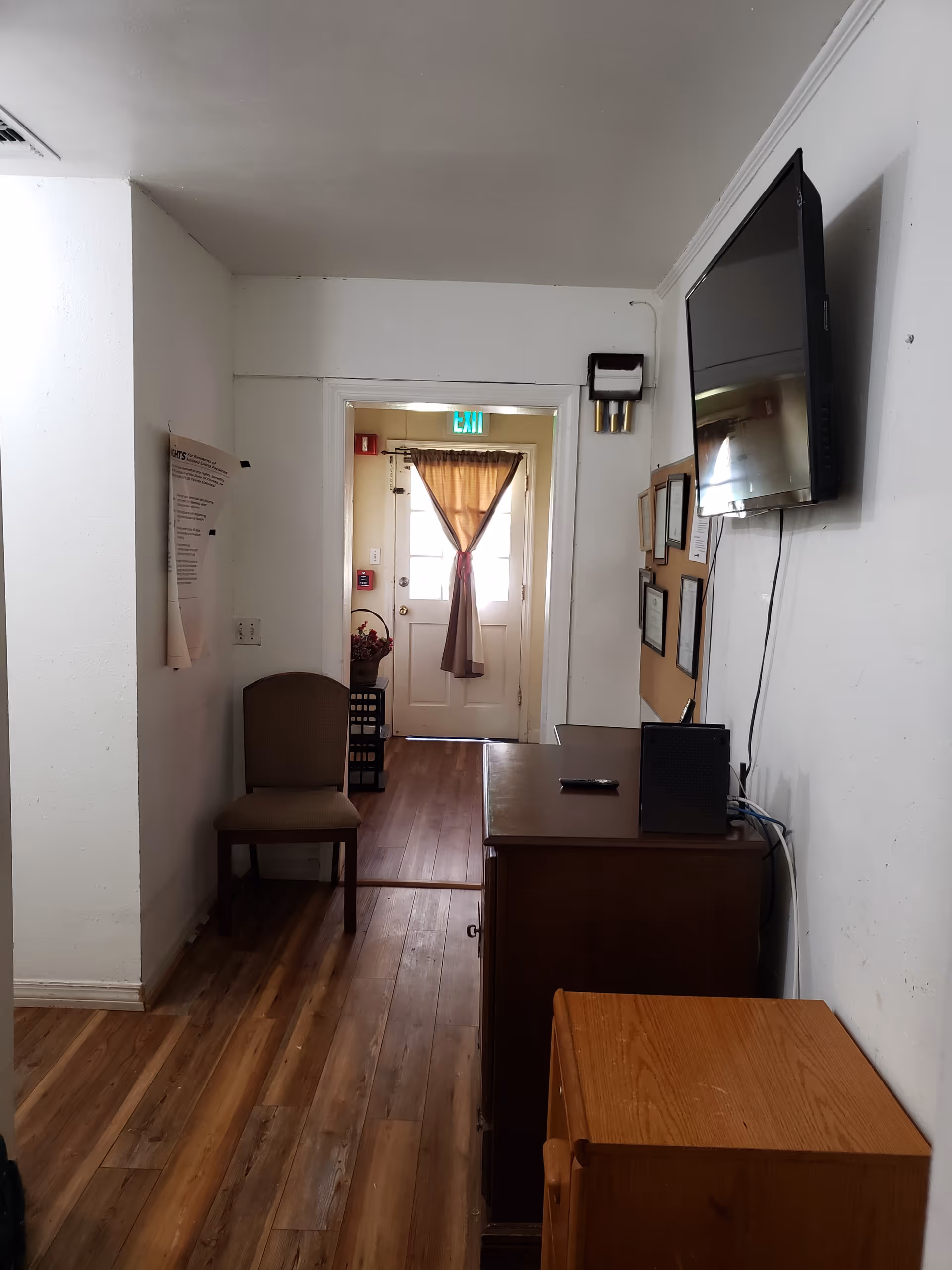 A narrow hallway with wooden flooring leading to a door with a brown curtain tied in the middle. On the left side, there is a single chair against the wall with a paper posted above it. On the right side, there is a wooden cabinet with a black speaker and a remote on top, a mounted flat-screen TV above it, and a bulletin board with framed documents. The walls are white and the area is lit by natural light from the door.