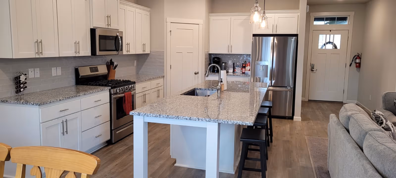 Modern kitchen with white cabinets, granite countertops, a kitchen island with a sink and three black stools, stainless steel appliances including a refrigerator, microwave, and stove, and a view into a living area with a gray couch and a front door in the background.