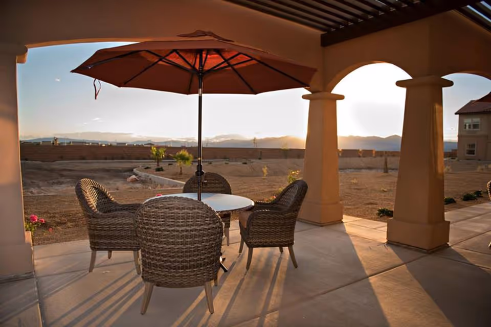 Covered outdoor patio with wicker chairs around a table and a large umbrella overlooking a desert landscape and distant mountains at sunset.