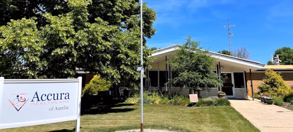 Exterior view of Accura HealthCare of Aurelia facility with a white sign displaying the facility's name in front of a well-maintained lawn, trees, and a building entrance under a clear blue sky.