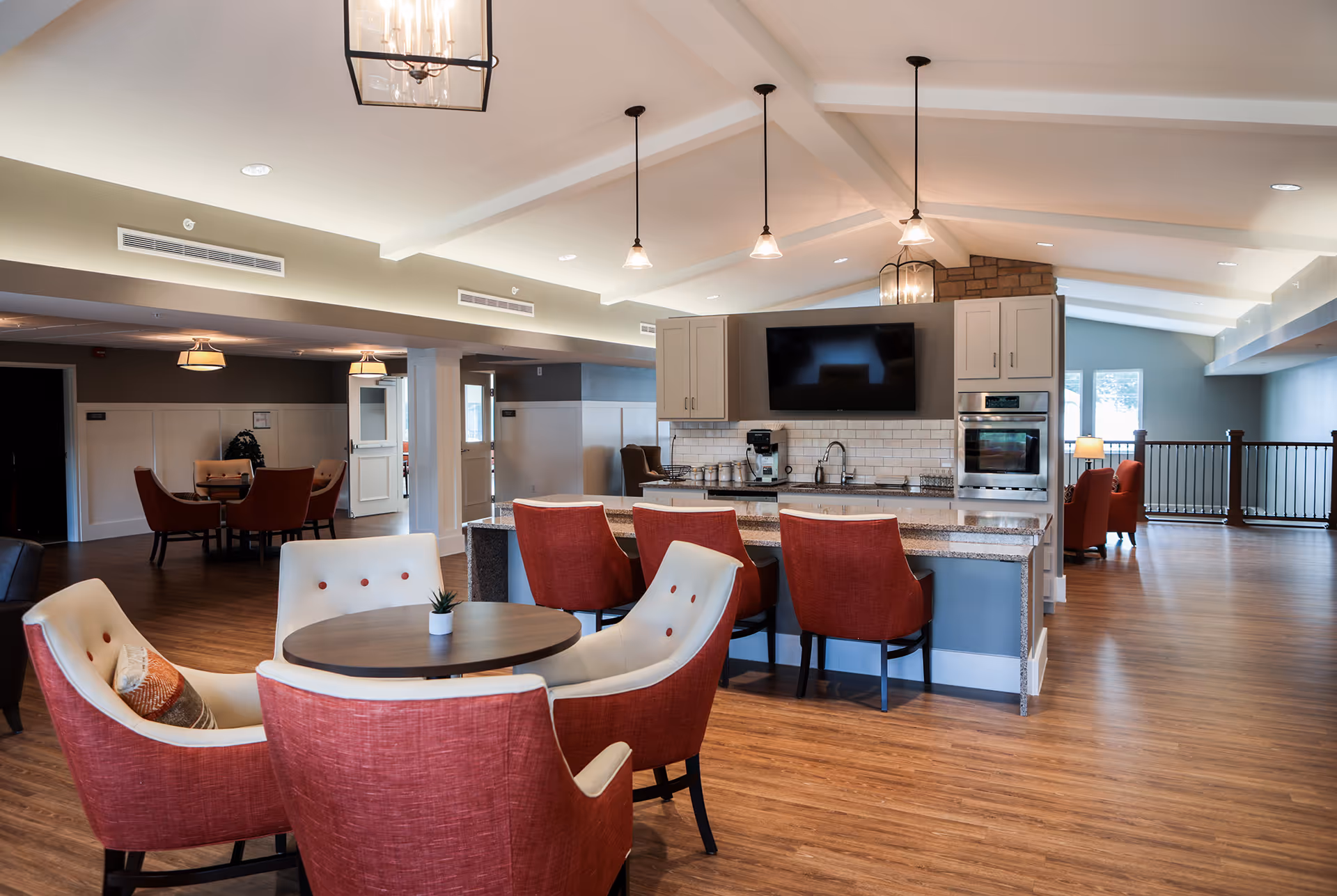 A spacious and modern common area in a senior living facility featuring a kitchen with granite countertops, white cabinets, a wall-mounted TV, and stainless steel appliances. There are red and white cushioned chairs around a small round table in the foreground and a kitchen island with red chairs. The room has wood flooring, pendant lighting, and large windows allowing natural light.