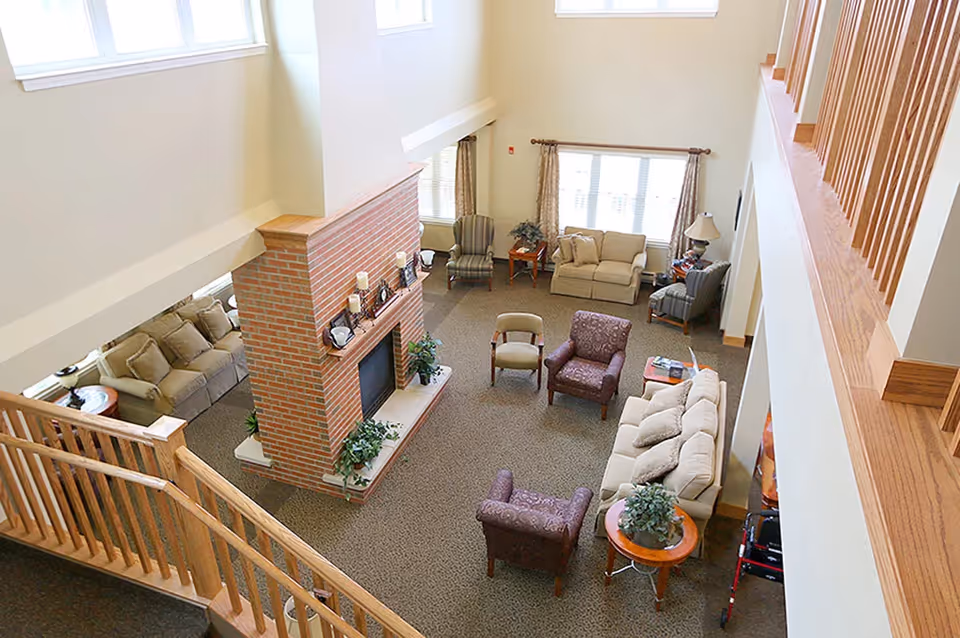 A spacious living room area with a high ceiling and large windows letting in natural light. The room features a central brick fireplace with candles and decorative items on the mantel. Surrounding the fireplace are multiple seating arrangements including beige and patterned armchairs, sofas, side tables with lamps, and potted plants. A wooden staircase with railings overlooks the room from above.