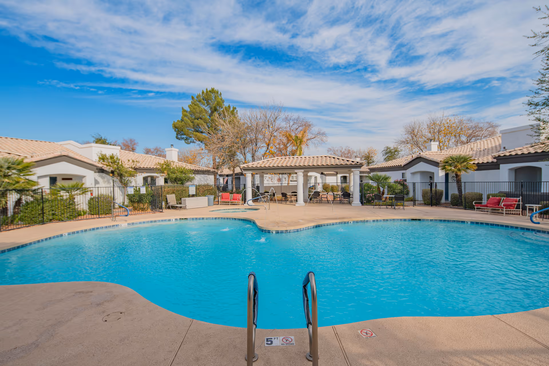 Outdoor swimming pool with clear blue water surrounded by a concrete deck, lounge chairs, and a shaded seating area with columns. The pool area is enclosed by a black metal fence, and single-story buildings with tiled roofs and trees are visible in the background under a partly cloudy sky.