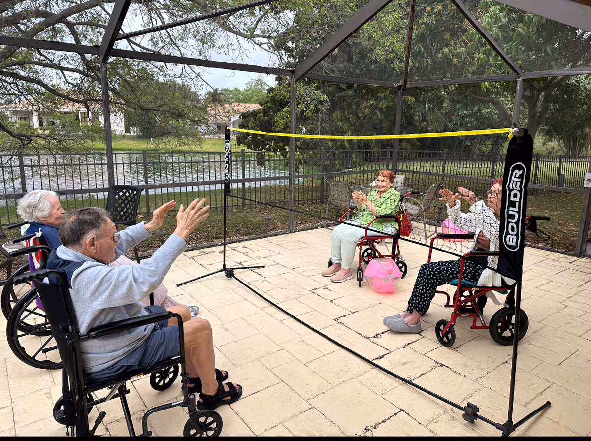Four elderly individuals in wheelchairs playing seated volleyball with a pink beach ball under a screened outdoor patio near a lake, surrounded by trees and a metal fence.