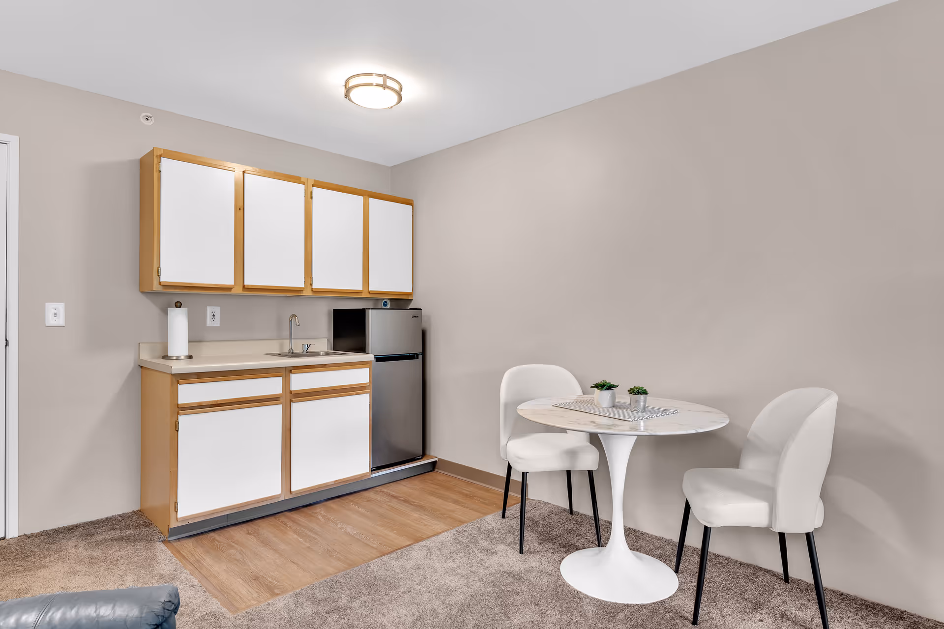 Small kitchenette area with wooden cabinets and a stainless steel mini refrigerator. Next to the kitchenette is a small round white table with two white upholstered chairs. The floor is carpeted except for the kitchenette area which has a wood-like floor. The walls are painted beige and there is a ceiling light fixture above.