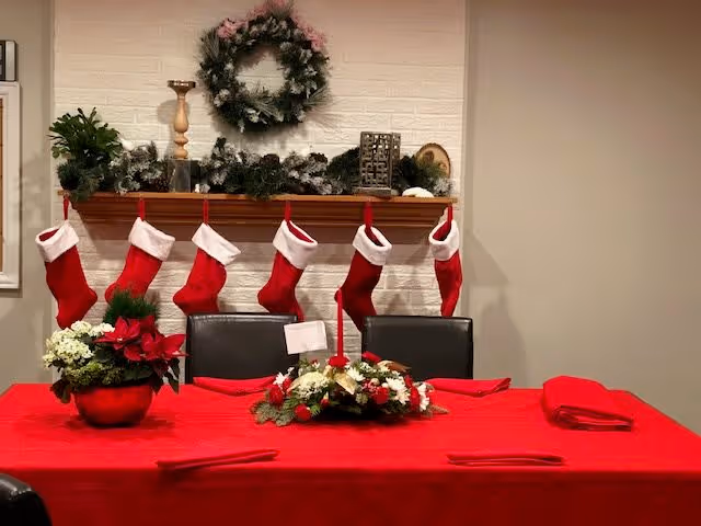A dining table covered with a red tablecloth and decorated with Christmas-themed floral arrangements. Behind the table, a wooden mantel is adorned with six red and white Christmas stockings, greenery, and a wreath hanging on a white brick wall.