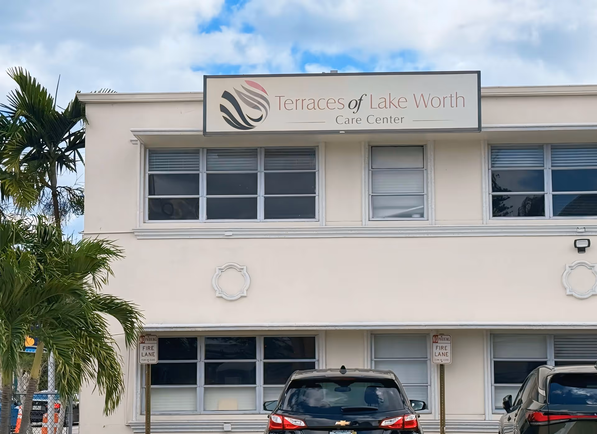 Exterior view of a two-story building with a sign that reads 'Terraces of Lake Worth Care Center' mounted above the second-floor windows. There are palm trees on the left side and parked cars in front of the building. The sky is partly cloudy.