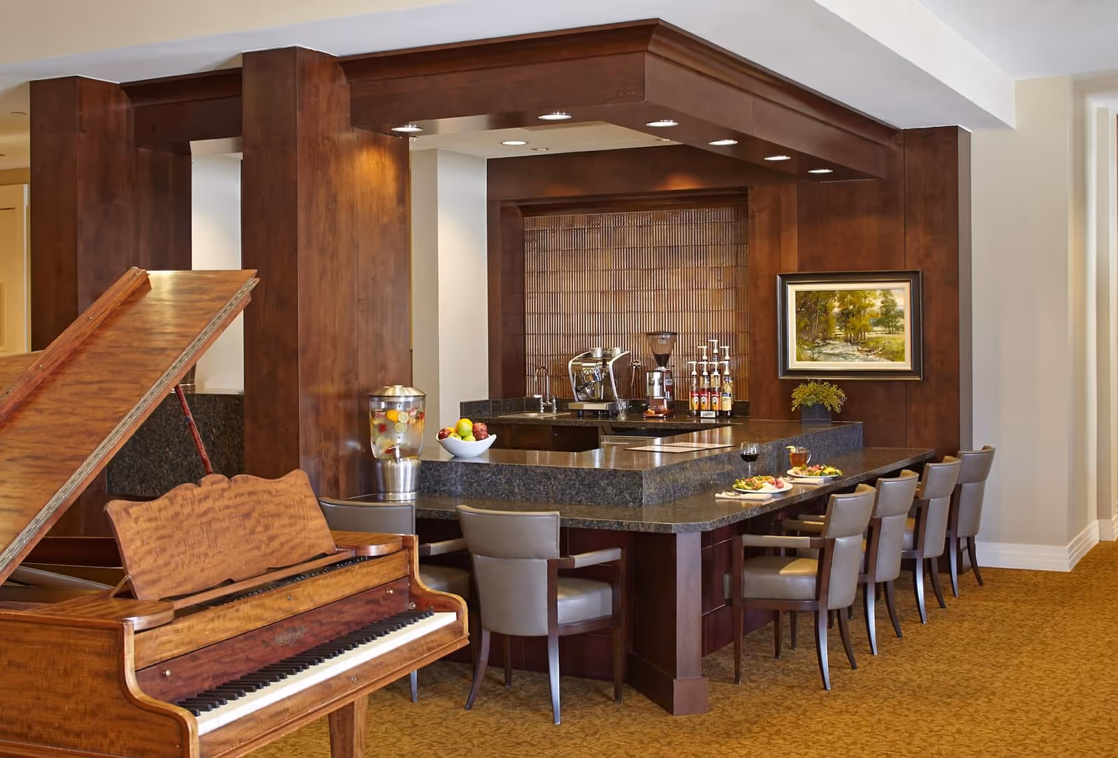 Interior view of a senior living facility lounge area featuring a wooden grand piano in the foreground and a granite countertop bar with several chairs. The bar area includes a coffee machine, syrup bottles, a water dispenser with fruit, and a bowl of apples. A framed painting and a small plant decorate the wall behind the bar.