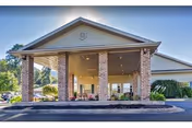 Front exterior view of a senior living facility with a large covered entrance supported by brick columns, surrounded by greenery and a clear blue sky.