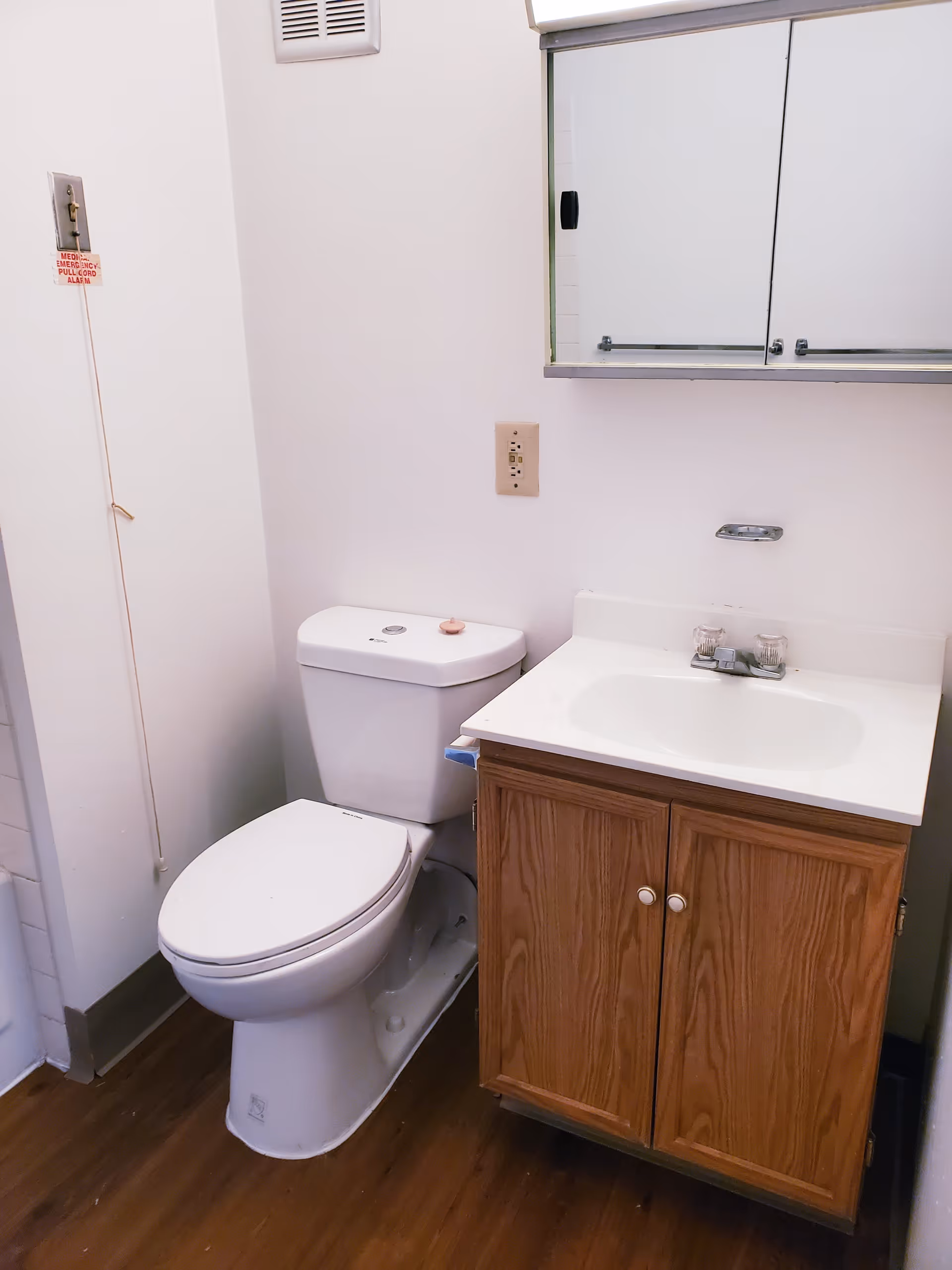 A small bathroom with a white toilet and a wooden vanity with a white sink. Above the sink is a mirrored medicine cabinet. There is a medical emergency pull cord on the wall next to the toilet.