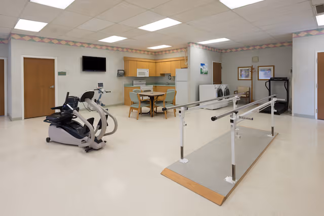 A rehabilitation room featuring exercise equipment including a recumbent bike and parallel bars for walking practice. There is a small kitchen area with a table and chairs, a microwave, refrigerator, and cabinets. A treadmill and a washer and dryer are visible in the background. The room has a tiled floor, white walls with a decorative border near the ceiling, and fluorescent lighting.