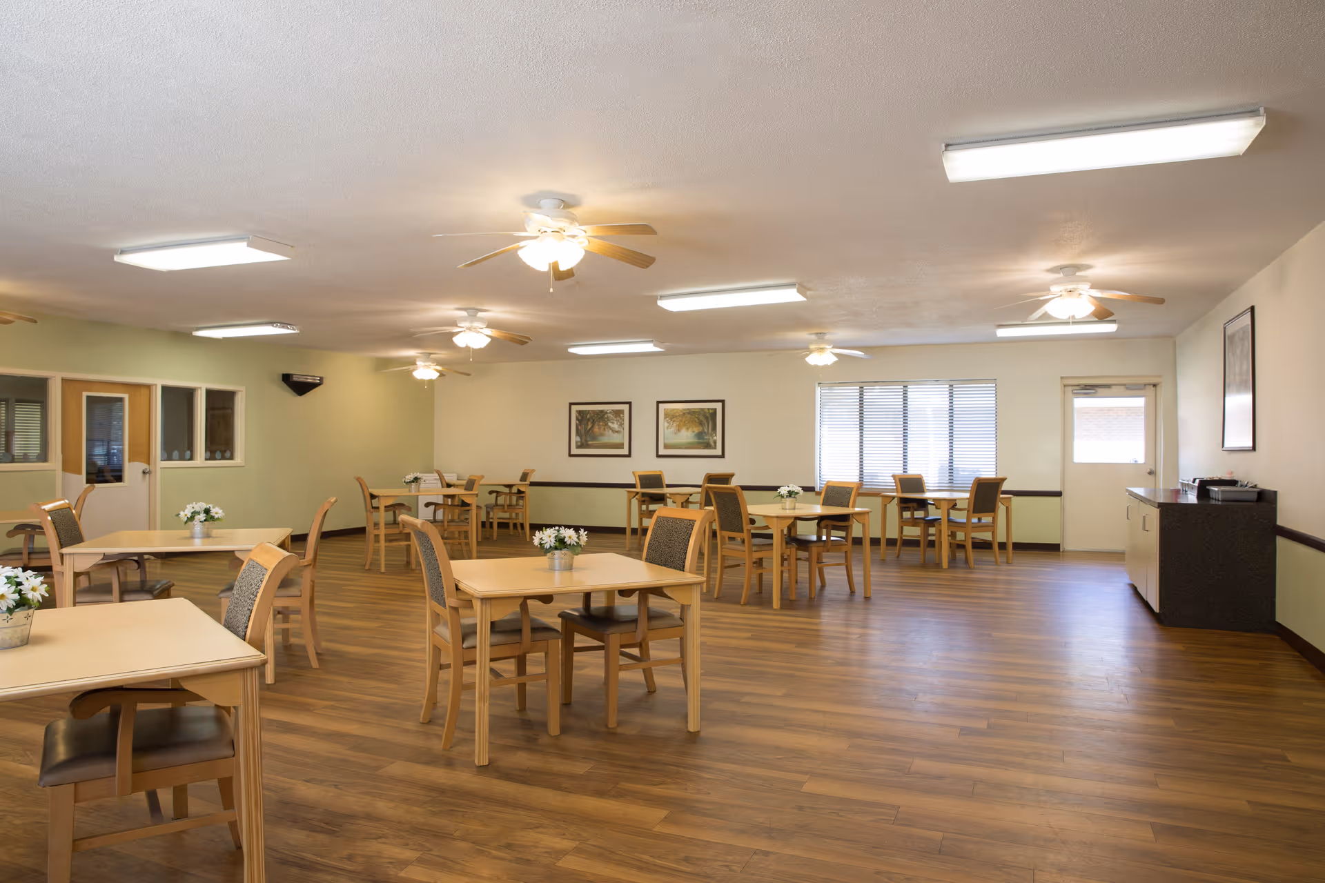 A spacious dining room with multiple wooden tables and chairs arranged neatly. Each table has a small flower arrangement. The room has wooden flooring, ceiling fans with lights, and fluorescent ceiling lights. There are framed pictures on the walls and windows with blinds letting in natural light.