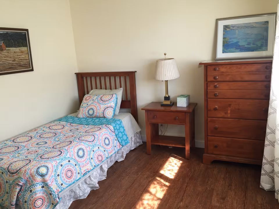A small bedroom with a single bed dressed in a colorful patterned quilt, a wooden nightstand with a lamp and tissue box, and a tall wooden dresser on hardwood floors.