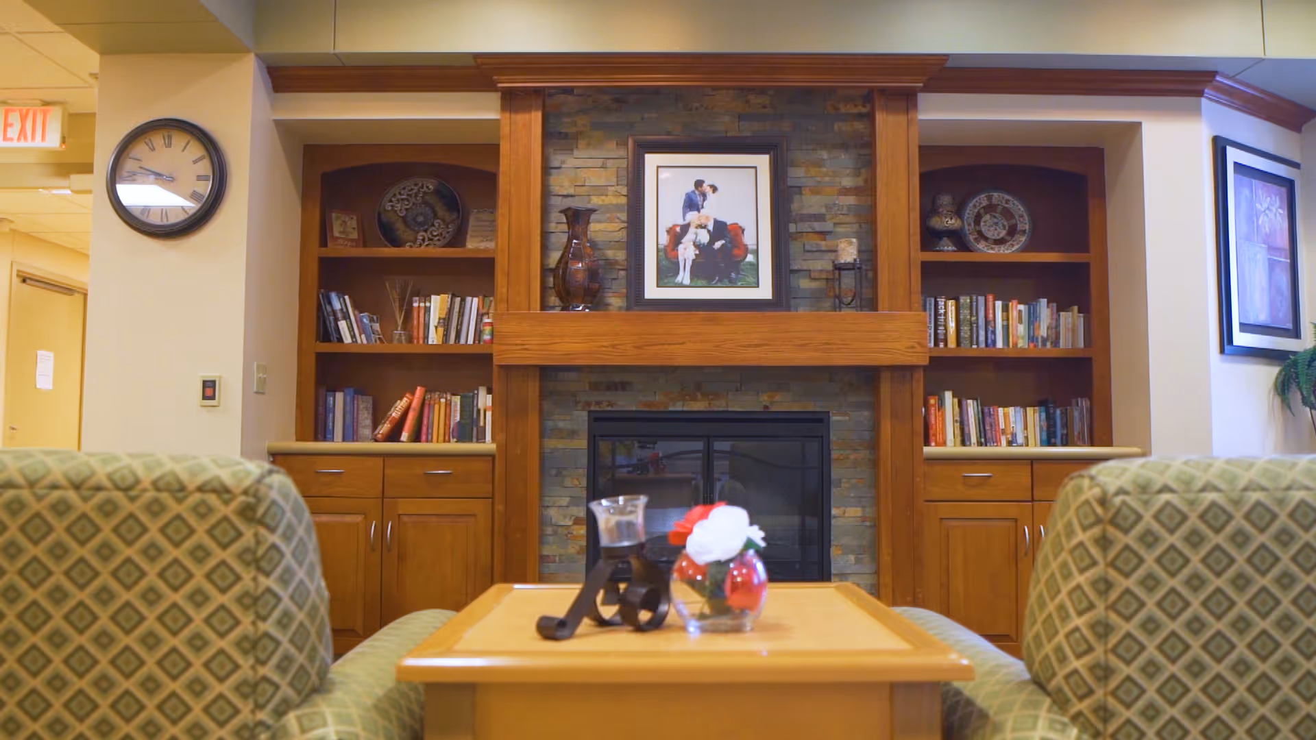 A cozy living room area in a senior living facility featuring a wooden fireplace with a stone backdrop. On either side of the fireplace are built-in wooden bookshelves filled with books and decorative items. In front of the fireplace is a wooden coffee table with a small glass vase containing red and white flowers and a candle holder. Two patterned armchairs face the fireplace. A clock is mounted on the wall to the left, and framed artwork is visible on the walls.