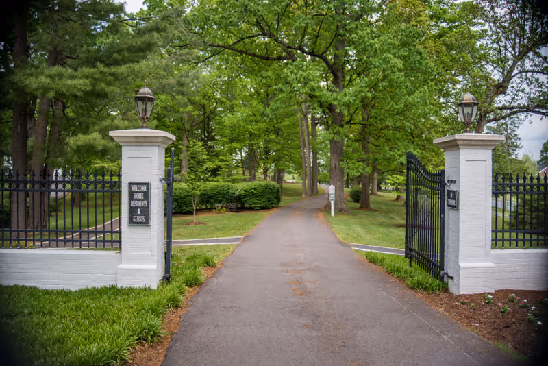 A paved driveway entrance flanked by white brick pillars with black wrought iron gates, leading into a green, tree-lined area with grass and bushes. Each pillar has a lantern on top, and one pillar has a sign that reads 'WELCOME HOME RESIDENTS & GUESTS'.