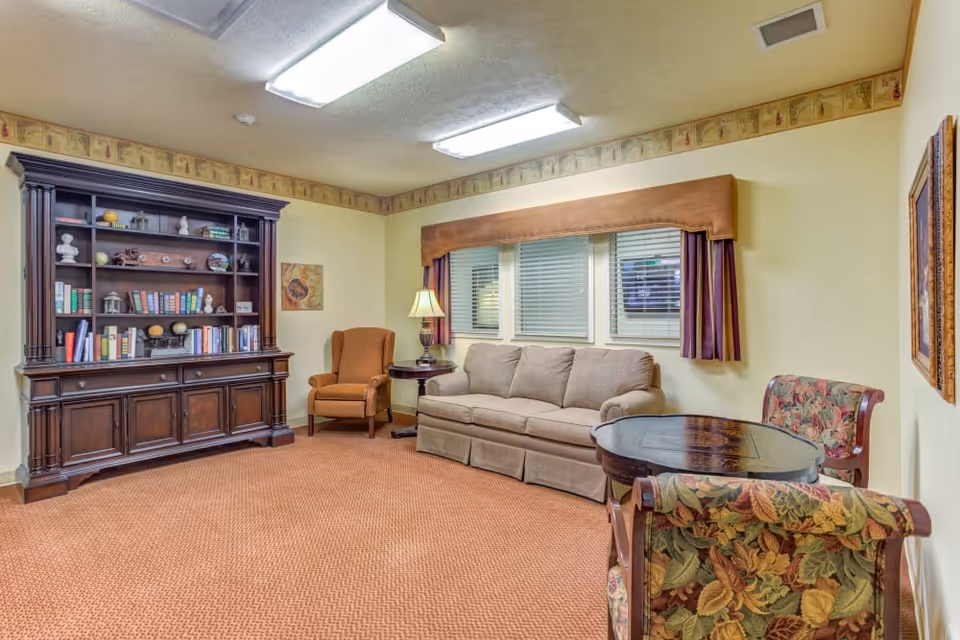 A cozy living-room style common area with a beige sofa, armchairs, a round table, lamp, and a large wooden bookcase.
