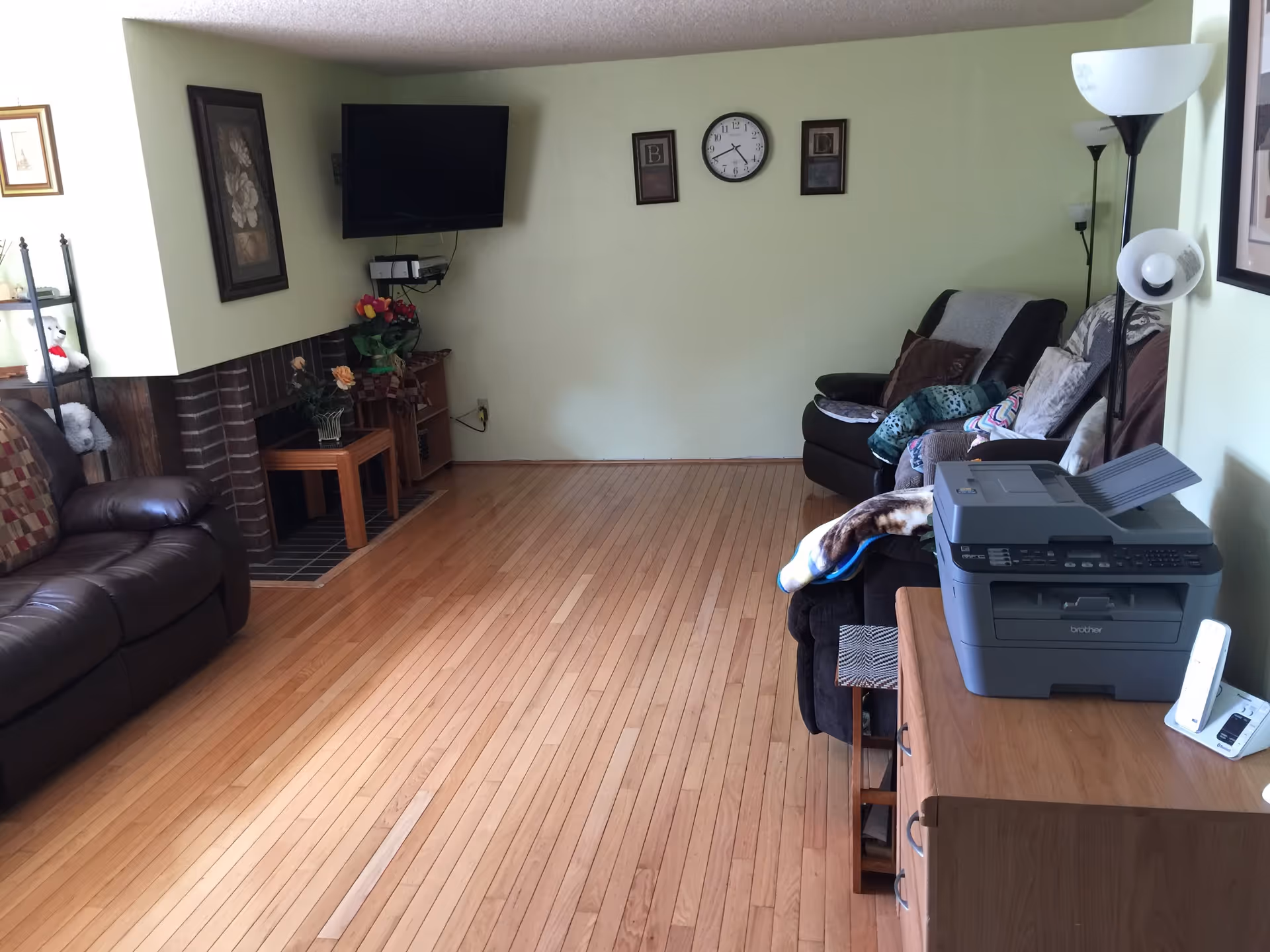 A living room with light green walls and wooden flooring. There is a brown leather sofa on the left and a dark recliner chair with pillows and blankets on the right. A flat-screen TV is mounted on the wall above a small wooden table with flowers. A clock and two framed pictures hang on the wall. A wooden dresser with a printer and a cordless phone is on the right side, next to a tall floor lamp.