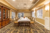 A formal dining room with a long rectangular table covered with a white tablecloth, surrounded by wooden chairs with beige cushions. The room has beige walls with white wainscoting, a patterned carpet, and a wooden china cabinet on the left side. There are windows on the right wall and a ceiling with recessed lighting.