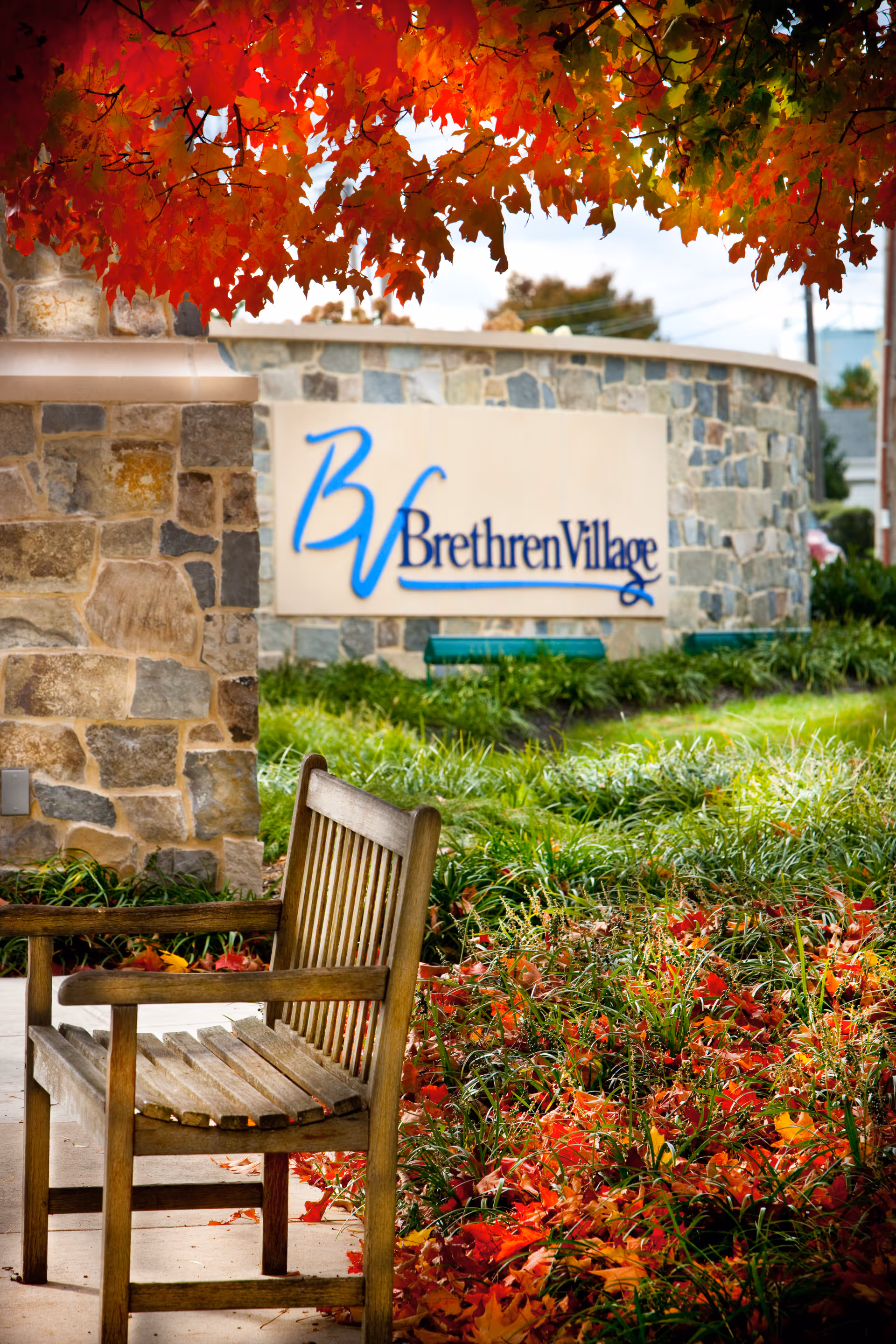 Wooden bench by landscaping and a stone entrance sign reading 'Brethren Village' amid autumn leaves.