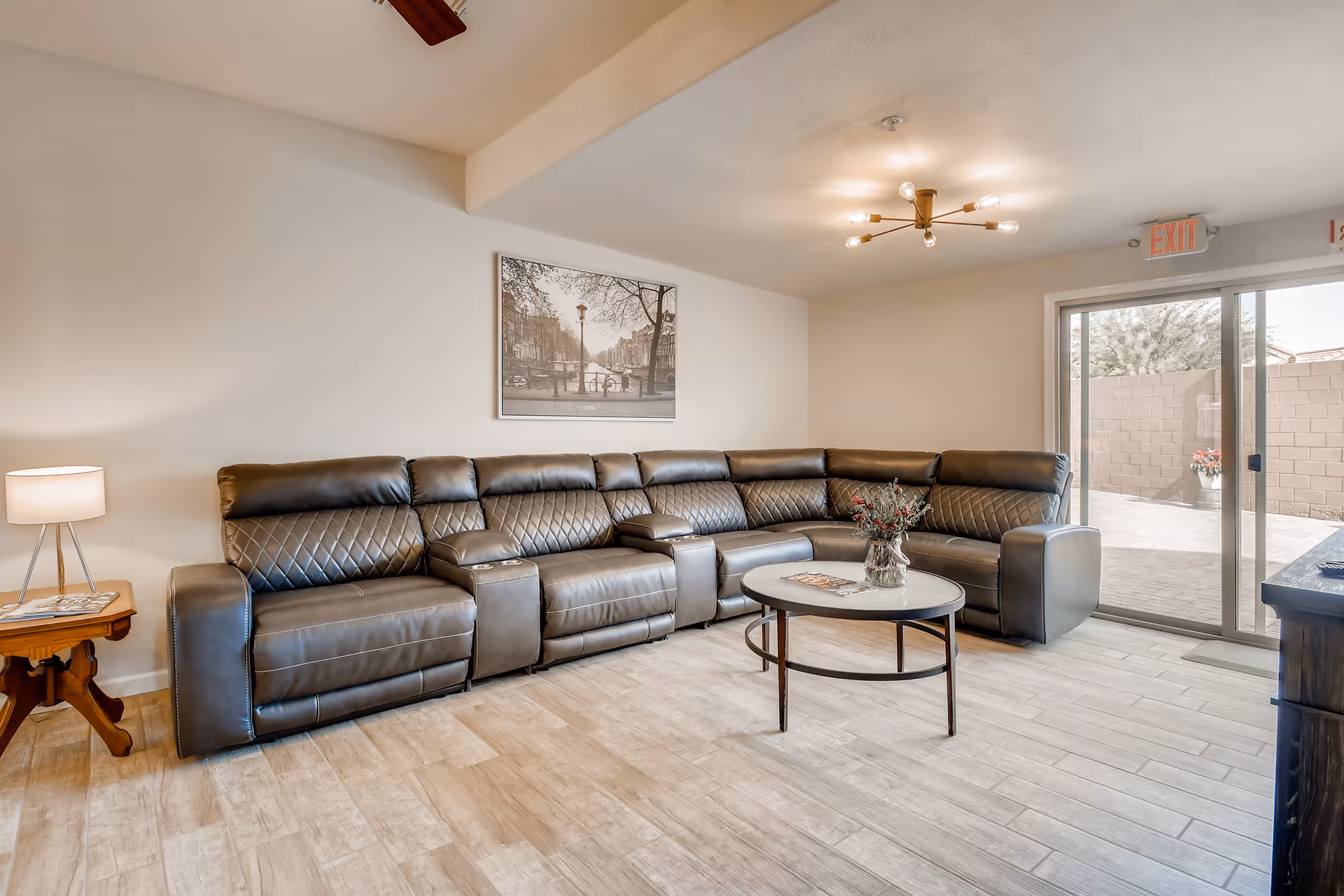 A spacious living room with a large dark brown leather sectional sofa arranged in an L-shape. A round coffee table with a vase of flowers sits in front of the sofa. There is a wooden side table with a lamp on the left side. A black and white framed picture hangs on the wall above the sofa. The room has light-colored wood flooring and a modern ceiling light fixture. Sliding glass doors on the right side lead to an outdoor patio area with a brick wall and some plants.