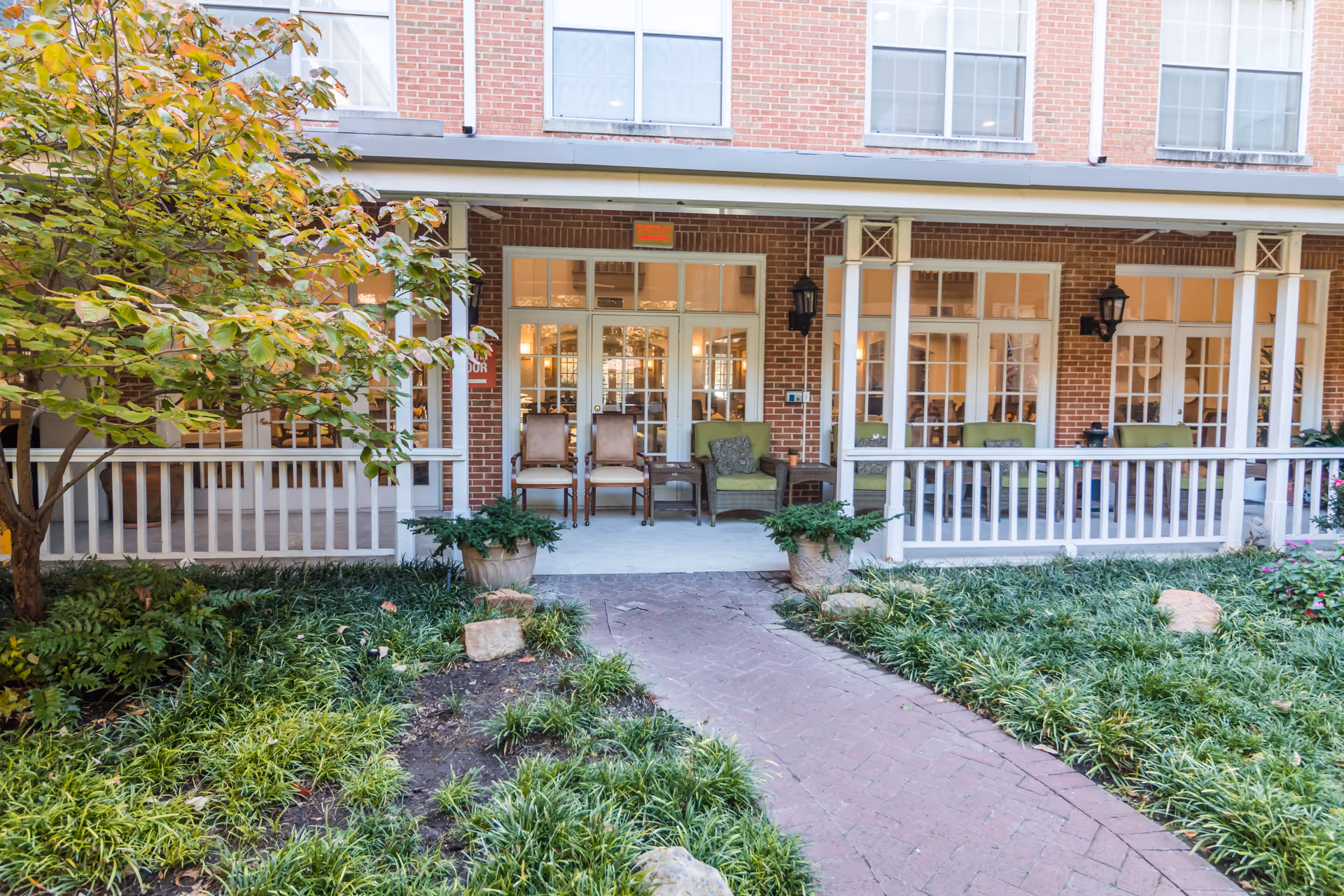 Front porch area of a brick building with white railings and columns. There are several chairs, including two wooden chairs and two green cushioned armchairs, arranged on the porch. A paved walkway leads up to the porch, surrounded by green plants and small trees.