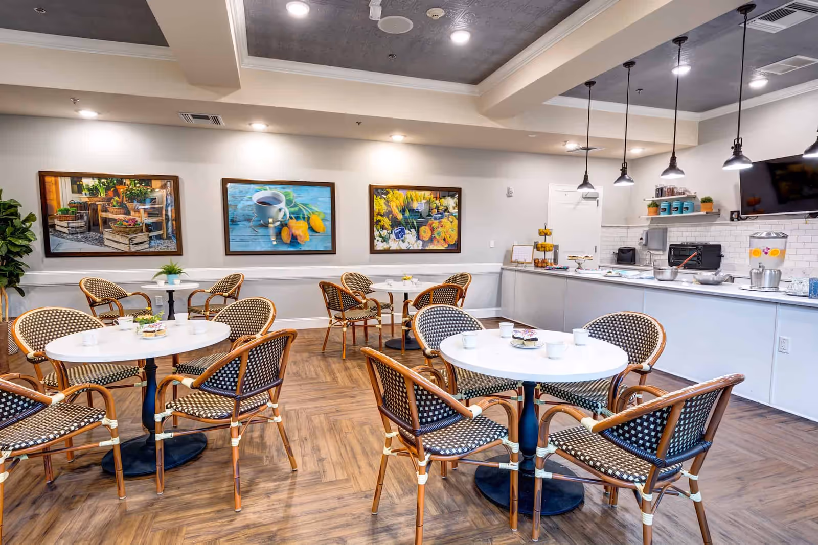 A bright and inviting dining area with round white tables and wicker chairs arranged neatly. The room features wooden flooring, three framed colorful paintings on the wall, and a counter with various kitchen appliances and refreshments. Pendant lights hang from the ceiling, illuminating the space.