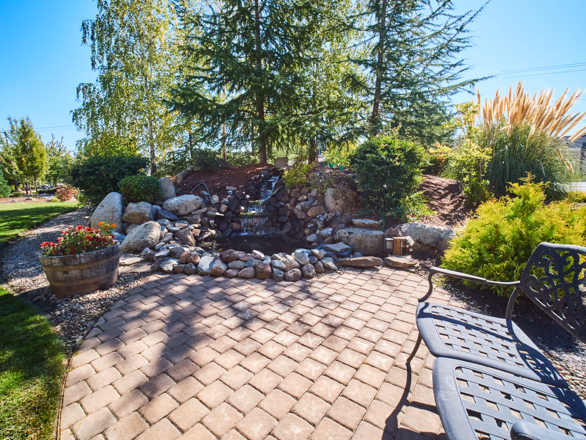 Sunlit outdoor patio with stone pavers, a metal bench, and a rock-lined pond with a small waterfall surrounded by shrubs and trees.