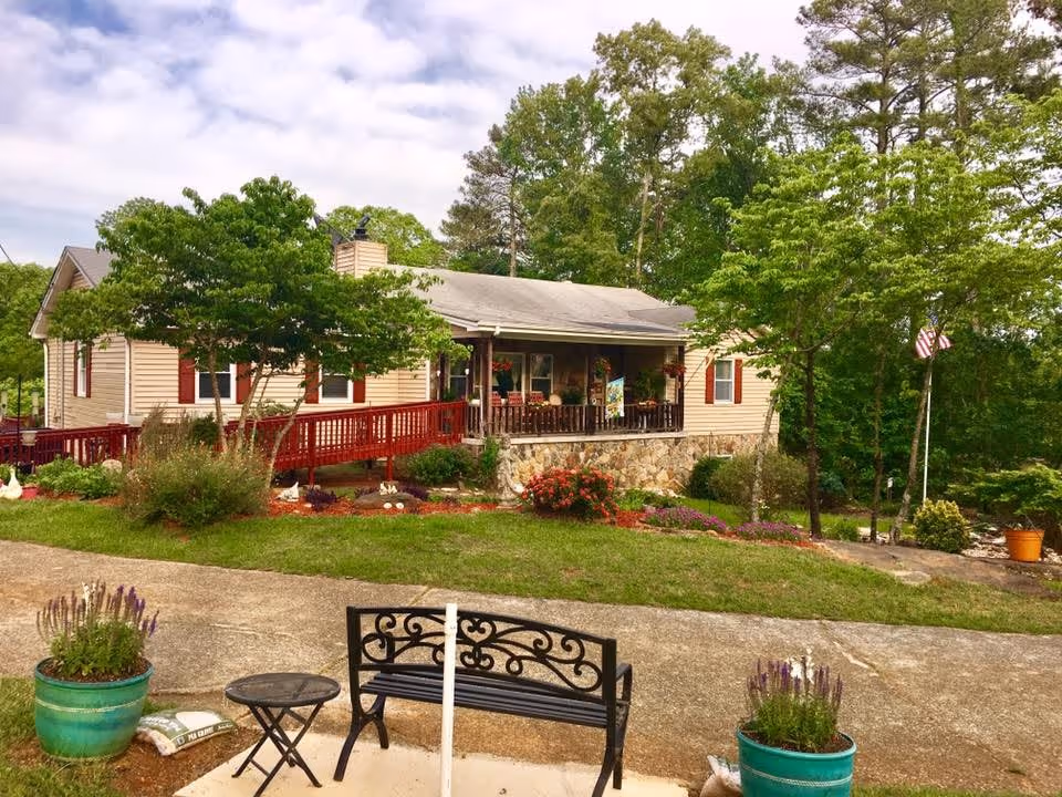 A single-story beige house with a stone foundation and a covered porch with hanging flower baskets. The house is surrounded by green trees and bushes, with a red wooden ramp leading to the porch. In the foreground, there is a black metal bench, a small round table, and two large green flower pots with purple flowers. The sky is partly cloudy.