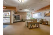 A cozy living room area in a senior care facility with beige carpeting, a stone fireplace, a large window letting in natural light, and comfortable seating including a sofa and armchairs. There are wooden side tables with decorative items and framed artwork on the walls.