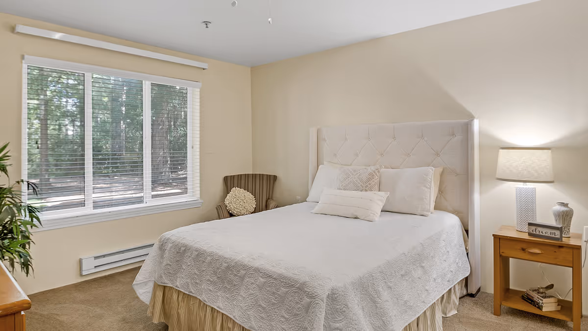 A cozy bedroom with a large window showing a view of trees outside. The room features a bed with a tufted headboard, white bedding, and several pillows. There is a wooden nightstand with a lamp, decorative items, and a small stack of books. A striped armchair with a round cushion is placed in the corner near the window.