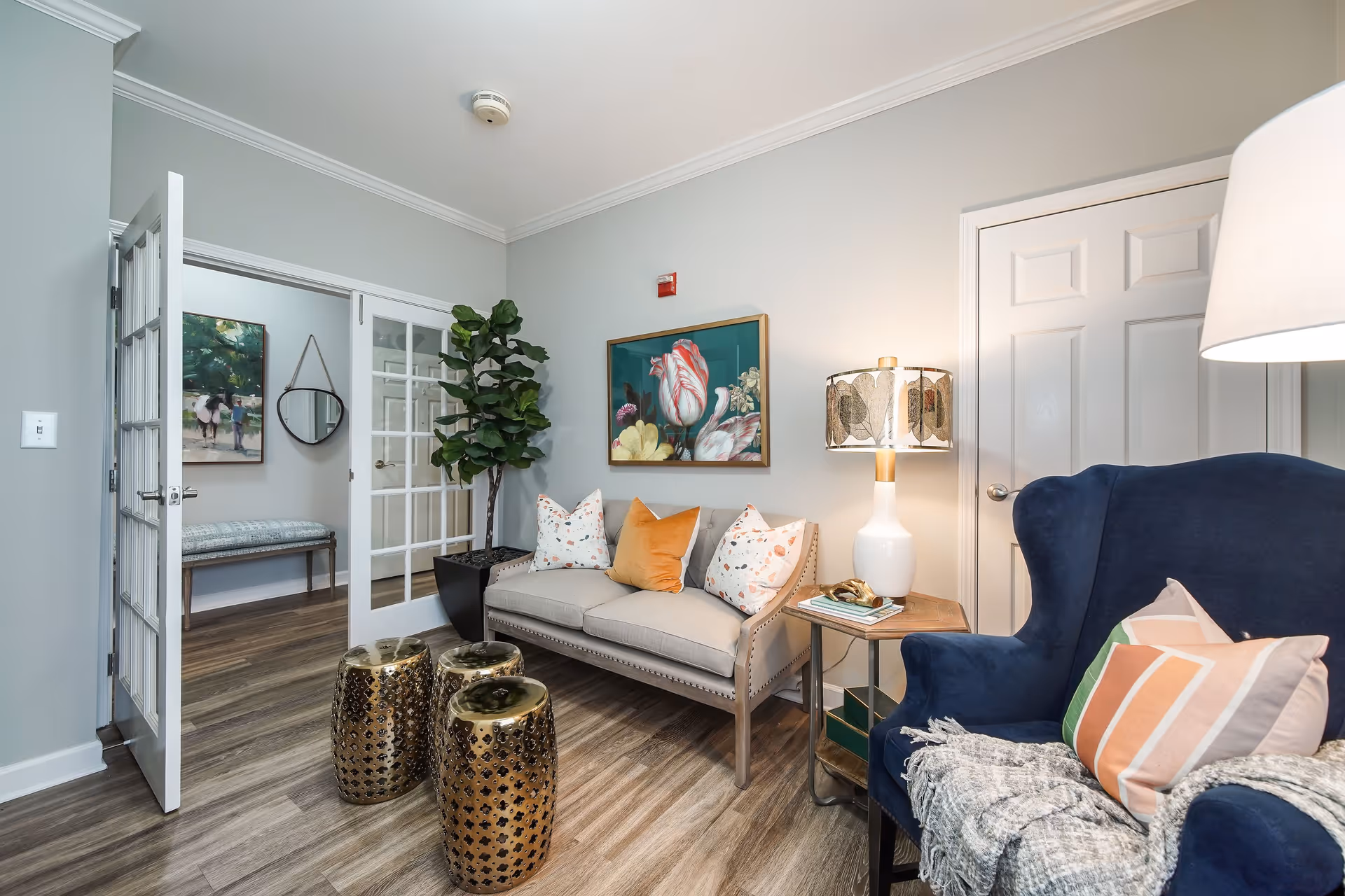 A cozy living room area with a beige loveseat adorned with three decorative pillows, a blue armchair with a striped pillow and a throw blanket, a wooden side table with a white lamp, and three gold decorative stools on a wood floor. There is a large potted plant next to the loveseat, a painting of flowers on the wall, and a door with white trim. In the background, a room with a bench, a round mirror, and a painting is visible through glass double doors.