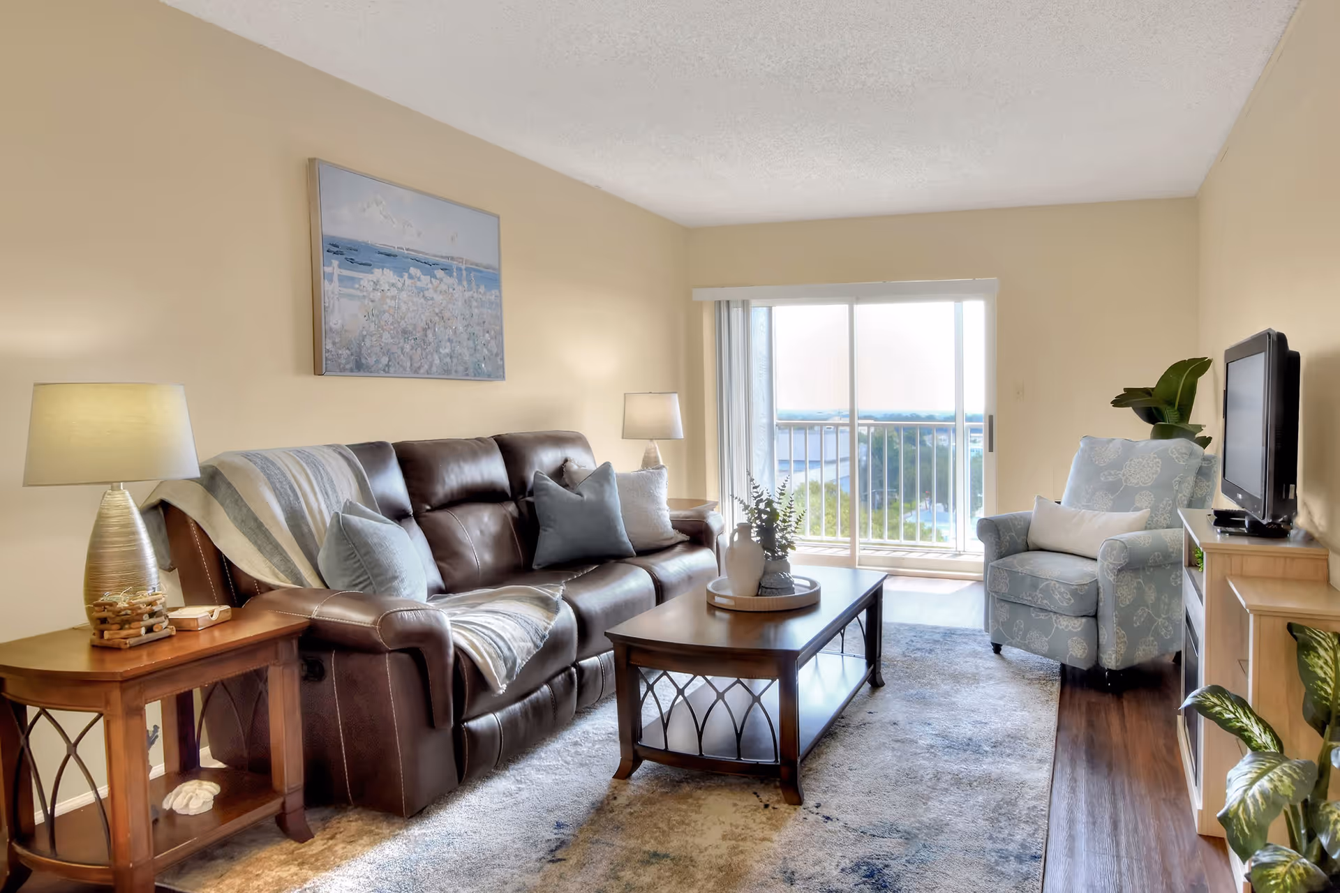 Bright living room with a leather sofa, armchair, coffee table, TV stand, and sliding glass doors leading to a balcony view.