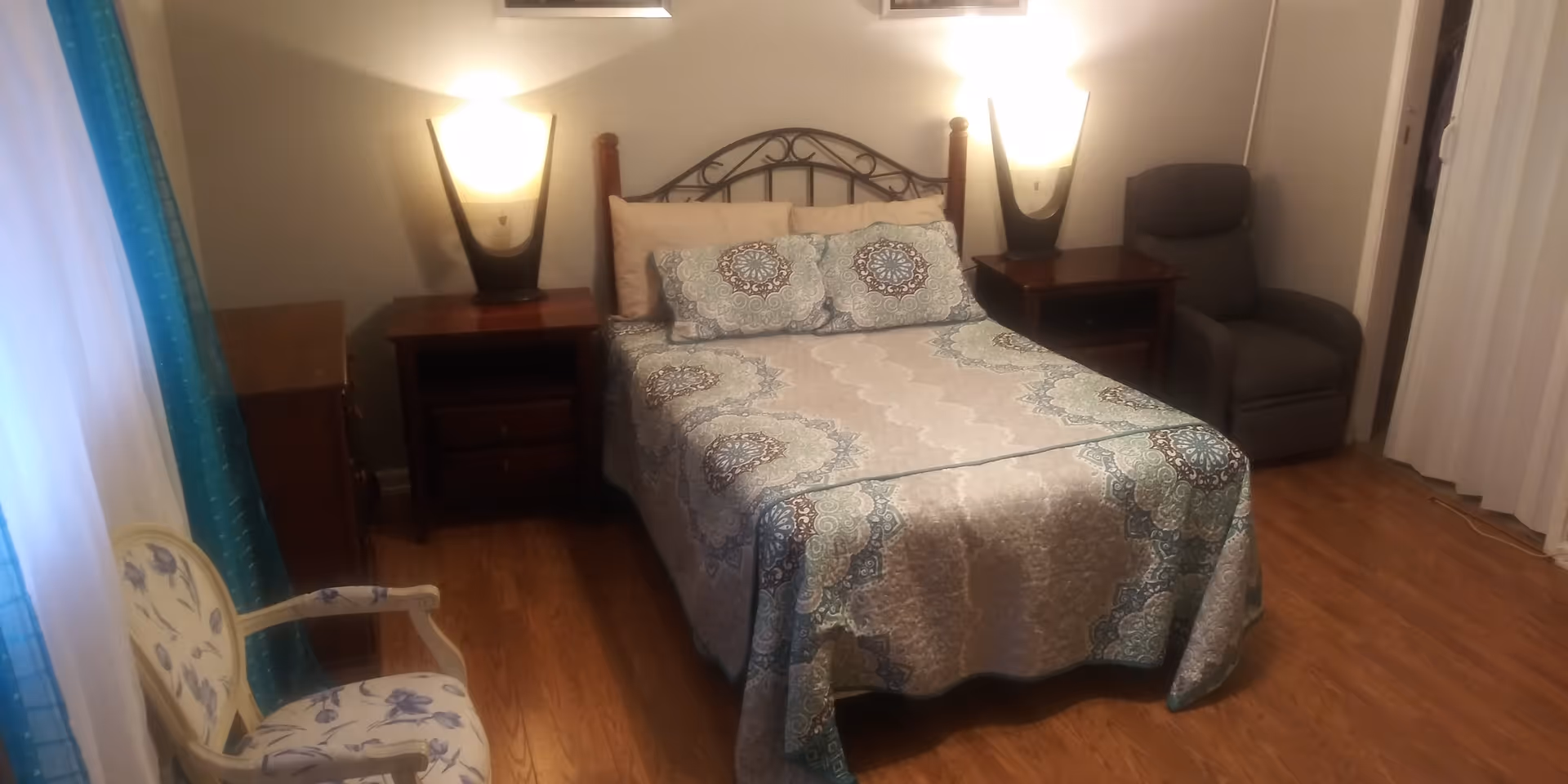 A bedroom with a double bed covered in a patterned bedspread and two matching pillows. There are two wooden nightstands on either side of the bed, each with a modern lamp. To the right of the bed is a gray recliner chair, and to the left is a floral upholstered armchair near a window with blue curtains. The floor is wooden, and the walls are painted light gray.