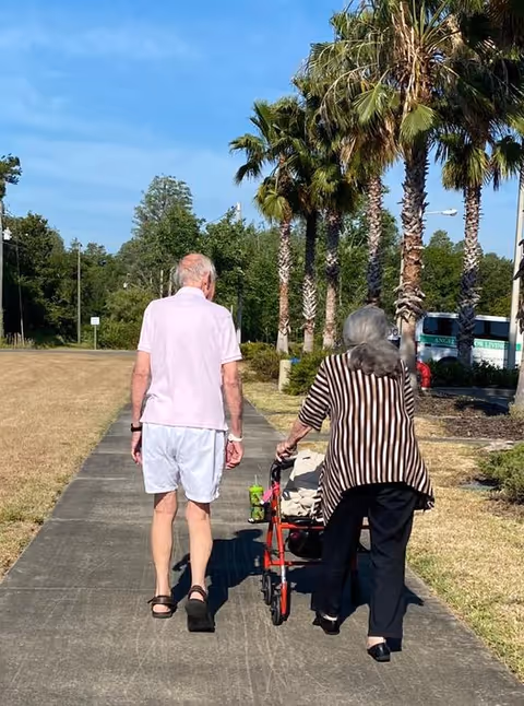 An elderly man and woman walking on a paved path outdoors. The man is wearing a light pink polo shirt and white shorts, while the woman is using a red walker and wearing a black and white striped top with black pants. Palm trees line the right side of the path, and there is a bus in the background with the text 'Angels Senior Living'.