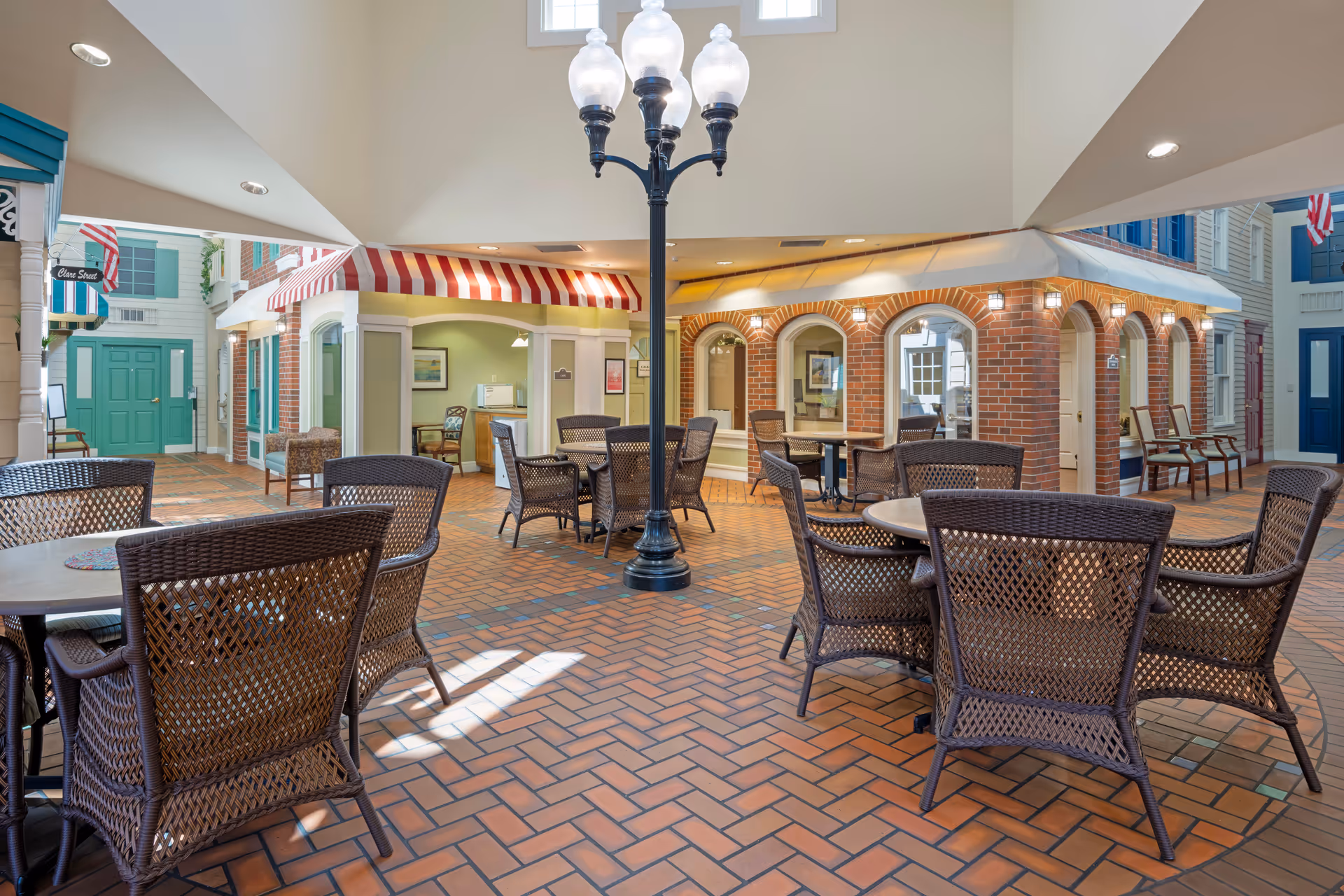 Indoor seating area with multiple round tables and wicker chairs arranged on a brick floor. The space resembles a small indoor village with storefront-style facades featuring awnings, windows, and doors. A tall black street lamp with five globe lights stands in the center. The ceiling is high with recessed lighting and skylights.