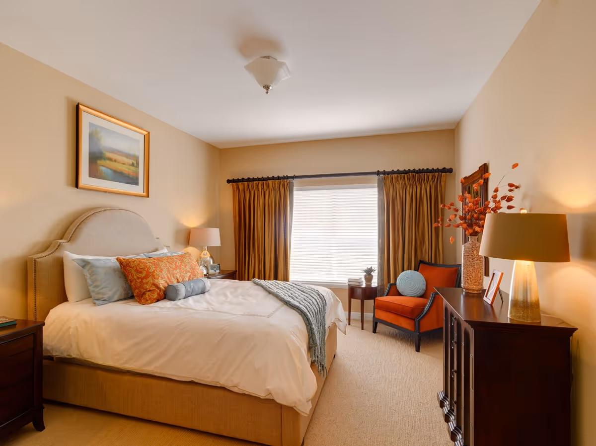 Well-lit furnished bedroom with a bed, bedside tables and lamps, an armchair by a window with drawn curtains and decorative accents.