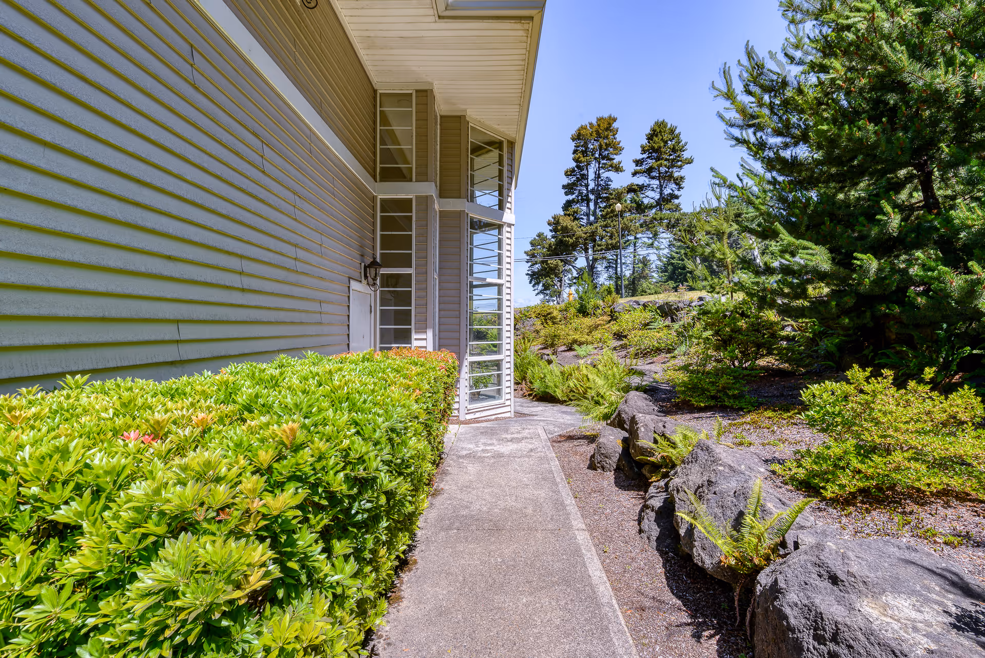Concrete walkway alongside a light-colored building with trimmed hedges and rocky landscaped greenery under a clear blue sky.